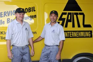 Zwei Mitarbeiter sind vor einem Dienstfahrzeug abgebildet. Zwei Mitarbeiter sind vor einem Dienstfahrzeug abgebildet.